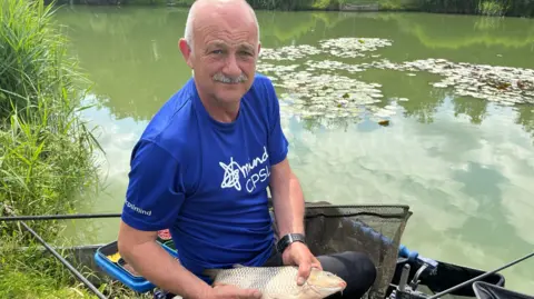 Jozef Hall/BBC Ian Darler holding a fish while sitting by a lake 