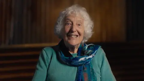 Gill Kirkham, pictured sat down in a teal top and blue scarf. She has short grey hair and is smiling at the camera
