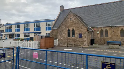 BBC Castel Primary School Guernsey. It two distinctly different buildings side by side, separated by a fenced playground area. n the right is an older stone building constructed from rough, brownish masonry. It has a steep, slate-grey roof and several narrow, arched windows. To the left is a much more modern two‑storey structure with a cream-colored exterior and bright blue window frames.