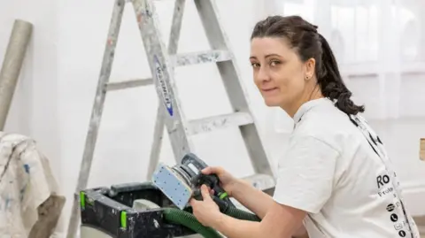 Rosie Russell Rosie Russell in work clothes, crouching down by a ladder and holding a sander