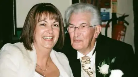 Lynda Costerd on the left with her father Geoff Toms on the right. They are attending a wedding. She has a fluffy white Faux fur shrug cardigan on, with a gold hanging necklace and brown bobbed hair with a fringe. He has a black suit jacket on with a gold tie and a cream flower in his pocket. Mr Toms is looking to the left of the camera and is smiling. He has glasses on.
