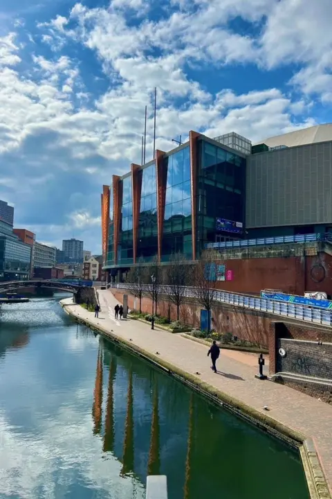 @BiteYourBrum A building with glass frontage and copper shingles stands alongside the canal. Part of it is reflected in the water. It is a sunny day with blue sky and a few white fluffy clouds.