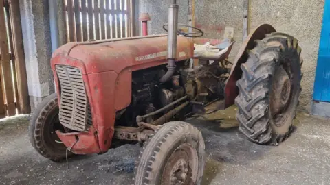Selkirk High School A rusty and dusty banged up old tractor sits in a garage. The front right tyre is deflated while the front grill is caved in.