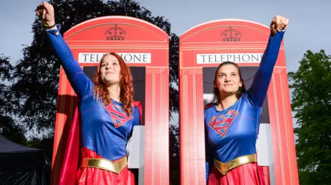 Two women wearing Superman costumes. Each raises one arm in the air in a superhero pose. They stand in front of red telephone boxes.
