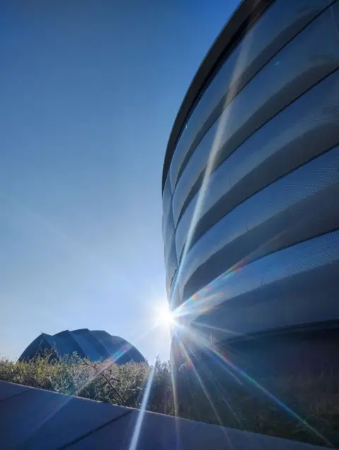 Donna Reekie A flare of sunlight inbetween two large concert arenas, with a blue sky