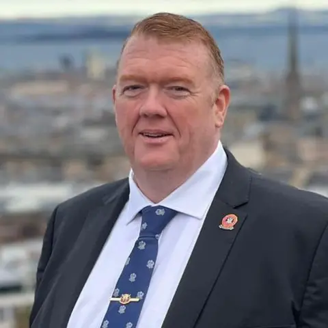 Scottish Conservatives A man with short hair in a blue suit and patterned tie looks directly at the camera. A town can be seen in the background.