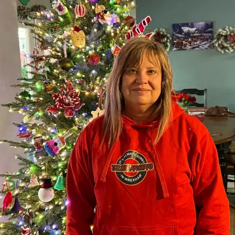 Jeff Ivey Janice in a red hoodie with a logo reading It's always sunny in Wrexham: The Declan Swans. She has blonde shoulder length hair and a fringe and is standing in front of a Christmas tree with white lights. 
