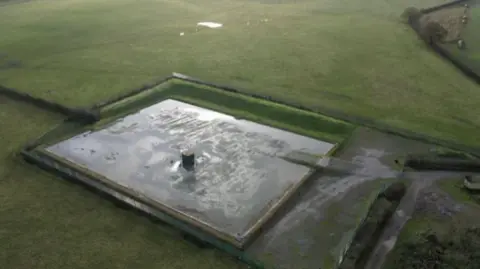 An aerial view of flat green fields with a rectangular watered surface in the middle. No people or vehicles can be seen.