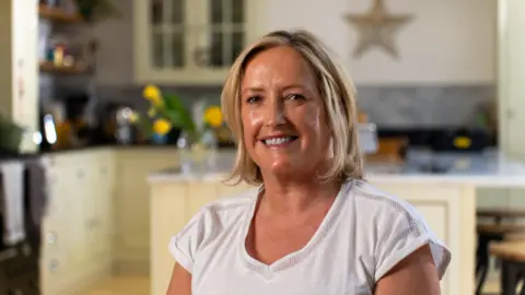 A woman sitting indoors in what appears to be a kitchen setting. She has short, light-coloured hair and is wearing a plain white short-sleeved top, positioned in the foreground and facing the camera. Behind the person, there are light-coloured cabinets, a countertop, and kitchen items such as jars, a kettle, and a vase containing yellow flowers, all slightly out of focus. The lighting is warm and soft, illuminating the person’s face while creating a gentle blur in the background.