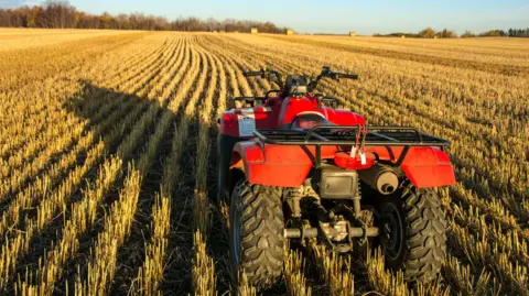 Getty Images A red quad bike in a freshly ploughed field. There are bales of hay in the background and the sun is shining.