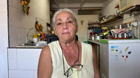 A woman wearing a yellow, short-sleeve top and with glasses on a string around her neck sits in front of a kitchen 