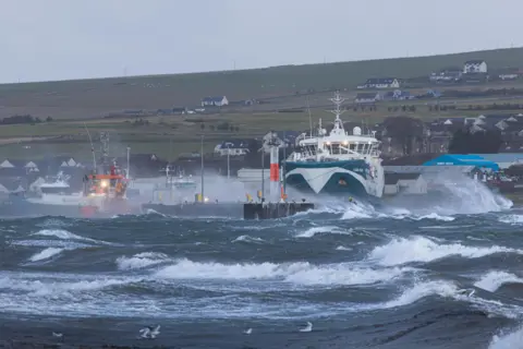 Mbdge/BBC Weather Watcher Choppy water at an island harbour. A blue and white ferry is berthed amongst the spray.