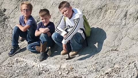The trio of fossil finders, Kaiden, Liam, and Jessin, pose in front of their find