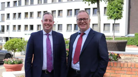 Department of Education Paul Givan wears a dark suit with a purple tie. Tom Bennett wears a dark suit with a red tie. Both have grey hair and Bennet is wearing glasses. They are standing outside in front of a white building,