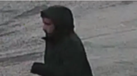 West Yorkshire Police A man whose face is partially obscured by a black hooded coat. The image is grainy.