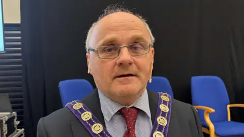 A man wearing glasses looks at the camera. He is wearing a suit with a red tie. Behind him are a number of blue chairs.