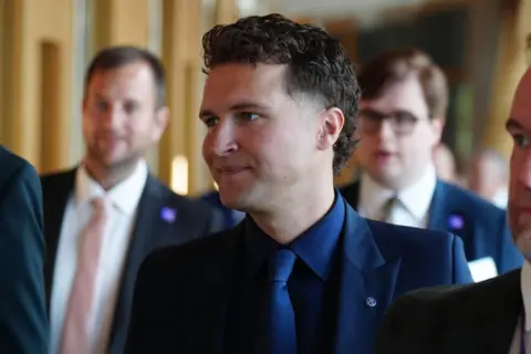 PA Media A man with curly black hair, in a dark business suit, with his head turned to the side 