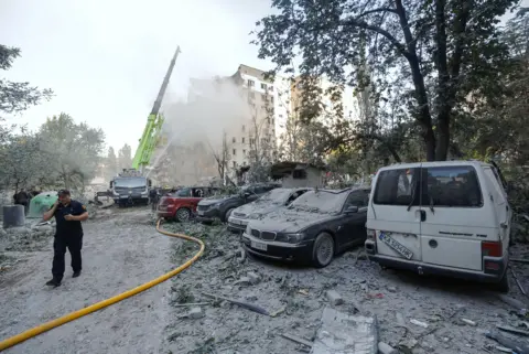 EPA A rescue worker wearing a navy blue uniform and cap is talking on the phone while walking past a row of five cars covered in dust and debris. In the distanct there is a crane and a hose shooting water pointing towards a building covered by a cloud of smoke and dust that has been partially damaged by a missile strike.