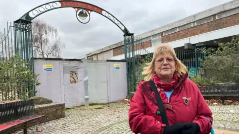 A woman in a red coat stands in front of boarded up gates