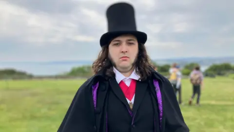 A young man dressed in austere Victorian costume and a top hat sneers at the camera