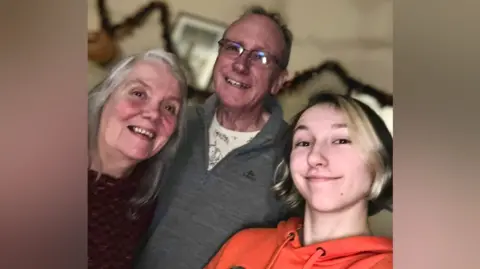 A selfie photo shows three people standing close together. There is a woman with grey hair and a purple jumper on the left, a man with grey hair and a grey sweater in the middle, and a person with short brown hair that has bleached blonde streaks on the right.