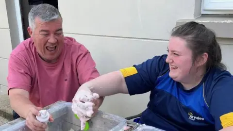 Conor and Ruby laughing. Conor has grey hair and is wearing a red t-shirt. Ruby has brown hair and is wearing a blue t-shirt.