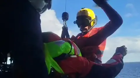 Row4Ukraine Two rowing crew members wearing life jackets and helmets being winched into a helicopter over the Atlantic with a rescuer helping them on board