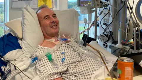 Ross Leslie, smiling at the camera from a hospital bed. He is wearing a hospital gown and is connected to equipment via tubes and wires.