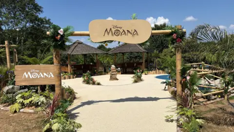 Jonny Michel A Moana themed garden with a Moana sign and lots of benches and greenery and two thatch-covered areas in the background.