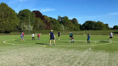 Two coaches dressed in navy sports attire spectate as a group of young hockey players dribble the ball