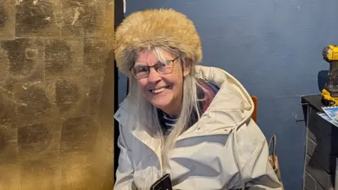 My Shrewsbury A woman with long grey hair, a pale coat and a fur hat in a room with one blue wall and one brick wall