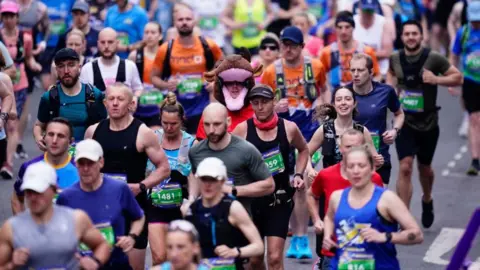 A large group of runners moving along a city street during a race. Participants are wearing colourful athletic gear, race bibs with numbers, and hydration packs. Among the crowd, one runner stands out wearing a full animal costume resembling a brown bull with horns and a pink snout.