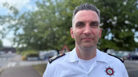 A man smiling at the camera. He has short, slicked-back, greying hair and is wearing a white shirt with black lapels on his shoulders. On the right-hand side of the shirt is a Lincolnshire Fire and Rescue badge.
