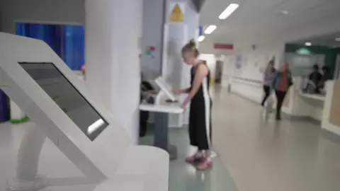 Tablets in reception at Royal Bournemouth Hospital Emergency Department. The screens are enclosed in white frames. The photo is out of focus in the background with a patient pressing the screen of one of the tablets. There is a corridor behind with white walls and floor with four figures standing. 