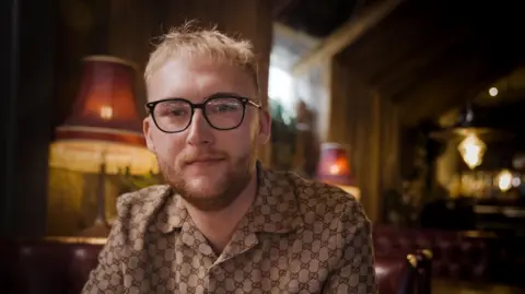 BBC/SpunGold TV Danny, a man with short blond hair and a beard, who wears a patterned shirt in neutral tones and black rimmed glasses, pictured in a room dimly lit by lamps.