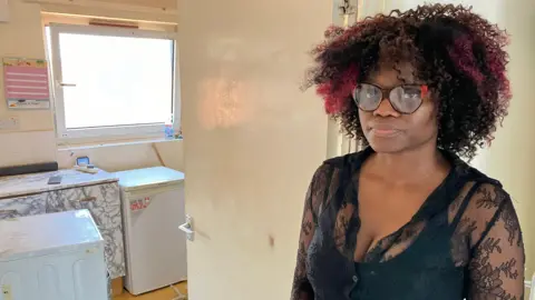 James Grant/BBC Woman with black curly hair and glasses stands in kitchen doorway