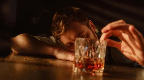 A man with brown hair and a beard leans his head on his arm on a table and gazes into a glass of whisky while running his index finger around the rim of the glass.