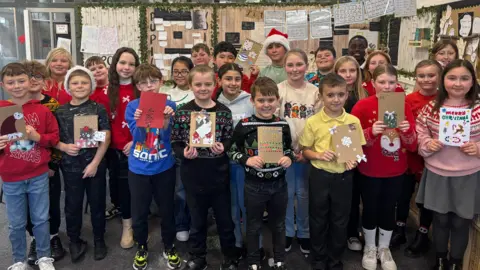 The image shows a group of children standing together in a classroom, holding handmade holiday cards. The classroom is decorated with festive elements, including greenery and hanging papers on the walls. Some children are wearing colorful sweaters with seasonal designs, such as snowflakes and characters. The cards they are holding feature various Christmas-themed decorations like snowmen, trees, and ornaments. In the background, there are posters and handwritten notes displayed on the wall, adding to the classroom setting.