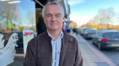 A man with short white and grey hair looks directly into the camera. He stands on pavement next to the glass front of a shop. Next to the pavement are several parked cars. The man is clean-shaven and wears a grey coat over a blue and dark blue striped shirt.