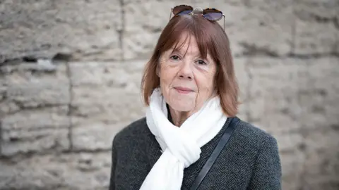 Getty Images Gisèle Pelicot, a French woman in her early seventies, wears sunglasses resting on top of her head, a white scarf around her neck, and a dark textured coat. The background features a stone wall, suggesting an outdoor or historic setting. Gisèle became internationally known after surviving nearly a decade of covert abuse by her husband, Dominique Pelicot. Between 2011 and 2020, Dominique drugged her and invited dozens of men to rape her while she was unconscious, filming the assaults.