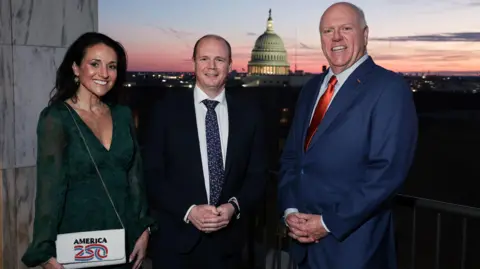 Three people in formal attire pose for a picture at sunset with Capitol Hill visible in the background. The woman on the left has black hair and is wearing a purse which says 'America 250'. The man in the middle is wearing a dark suit, the man to his right a blue suit.
