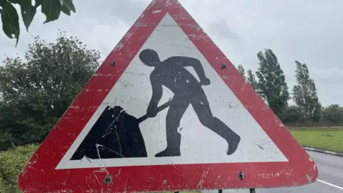 BBC Close up of a large roadworks sign displaying a silhouette of workman with a spade on a white background within a red triangle. Trees and other greenery can be seen behind it.