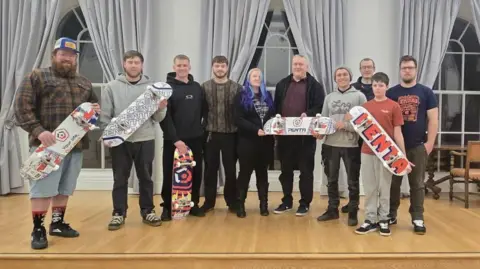 Corsham Skatepark Project Group The campaign group stand together in what appears to be a town hall. They are smiling, and some of the people are holding skateboards. 