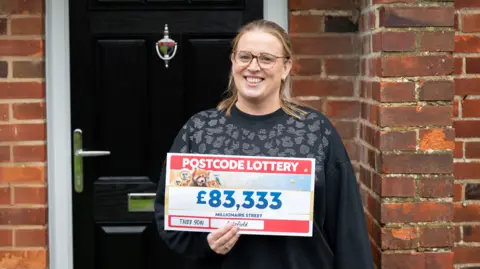 Mujer sonriendo frente a una puerta con un billete de lotería de £83,333
