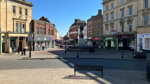 BBC A shot of Bridgwater High Street. In the foreground is a bench draped in a shadow, while further down the road is a statue. The pedestrianised area is flanked by a number of shops. There is a blue, sunny sky.