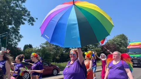 A person with brown hair tied back, wearing glasses and a purple top, holding a rainbow umbrella aloft. There are other people in purple tops alongside her. People dressed in red outfits are following in a parade behind. There is a lorry behind them.