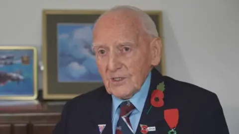 Bernard Morgan has short white hair. He wears a blue shirt with a burgundy and blue tie under a dark blue blazer. He wears medals, a pin badge poppy and a paper poppy on his collar. 