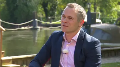 Tim Mcmahon has short hair and is wearing a pink shirt and a blue jacket. He is sat in front of the River Thames, which is flowing by in the sun. 