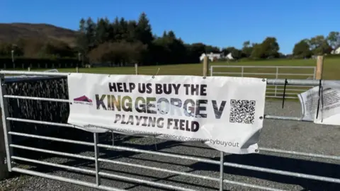 A white fabric sign attached to a metal farm-style gate. The sign has a message that reads: "Help us buy the King George V playing field.