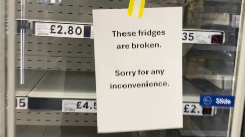 A sign saying 'these fridges are broken, sorry for any inconvenience'. There are empty fridge shelves behind it. 